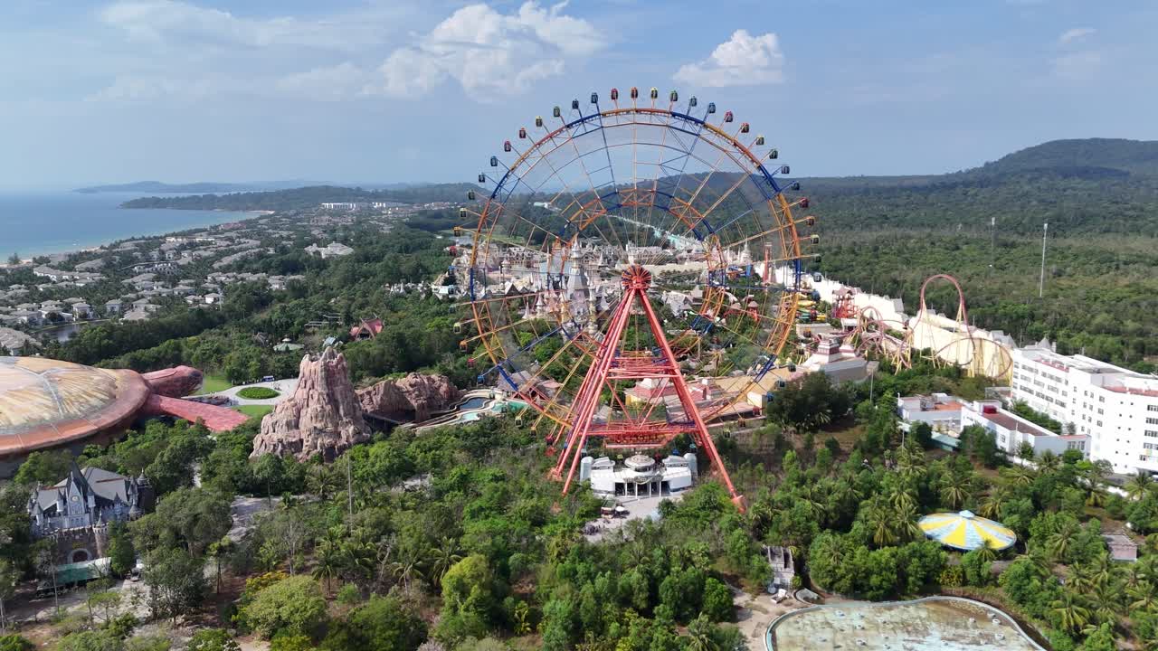 The aerial perspective amusement park, its colossal Ferris wheel offering panoramic views of the coastline and ocean. Teeming with themed structures and lush vegetation, the park has coastal hotels.