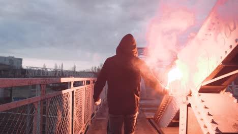 back view of a young man in hoodie and balaclava with red burning signal flare on the road under an old steel frame bridge, slow motion