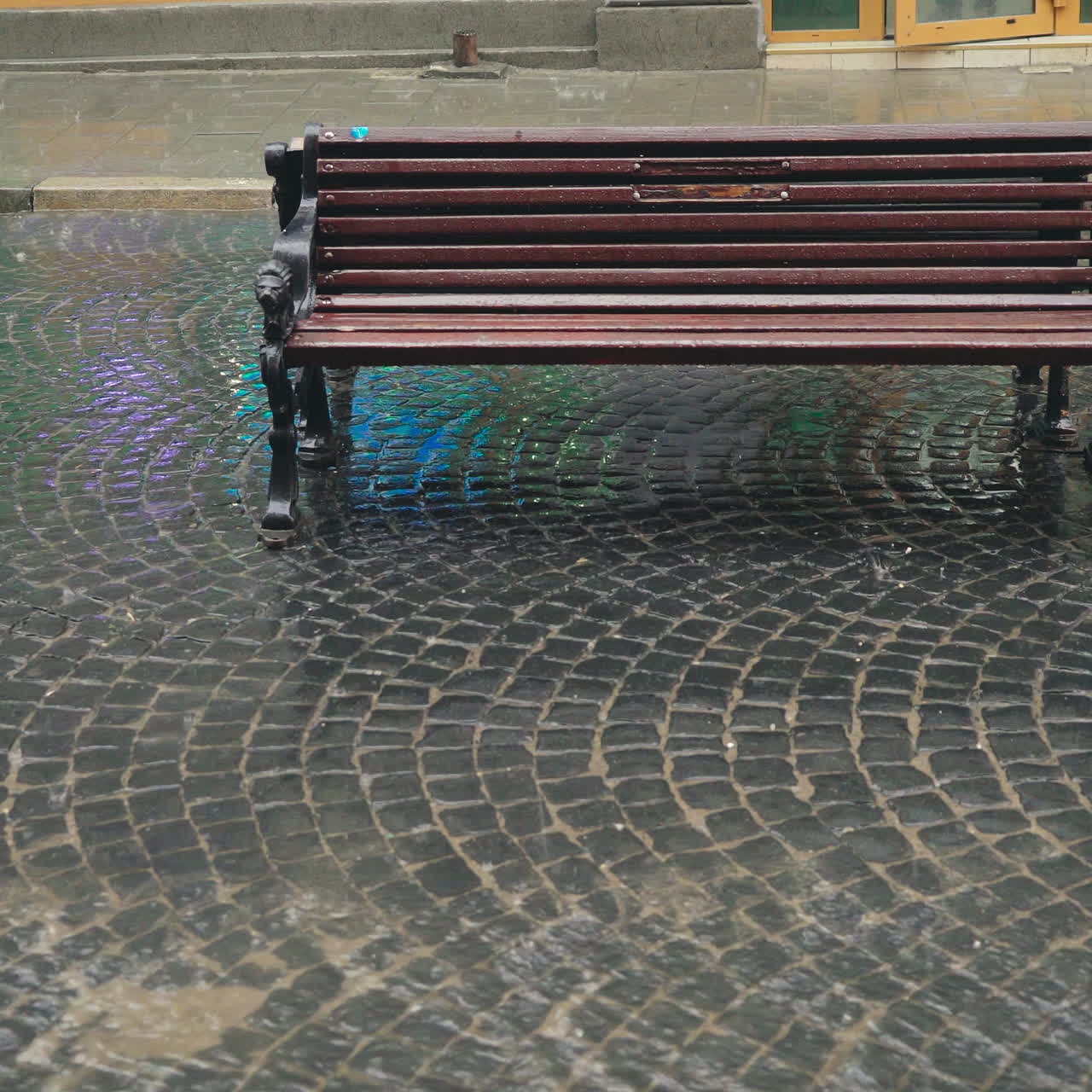 Heavy rain is falling on the pavement and modern wooden bench outdoors in the city. Stream of water are flowing through the pavement in a rainy weather.