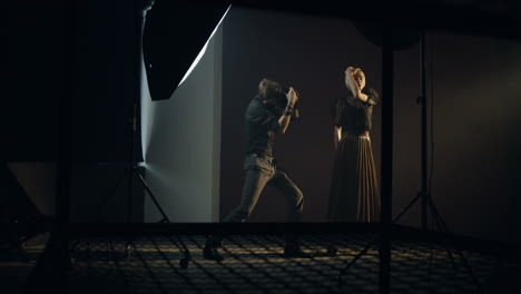 distant view of caucasian young female model posing in the dark studio while the photographer taking shots of her