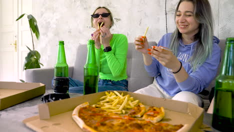 happy girl friends laughing and having fun with fries while sitting on sofa at home