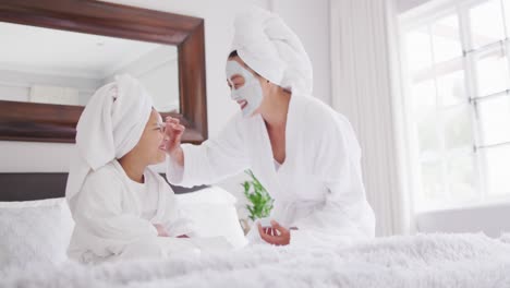 video of happy asian mother and daughter in robes moisturizing faces and having fun