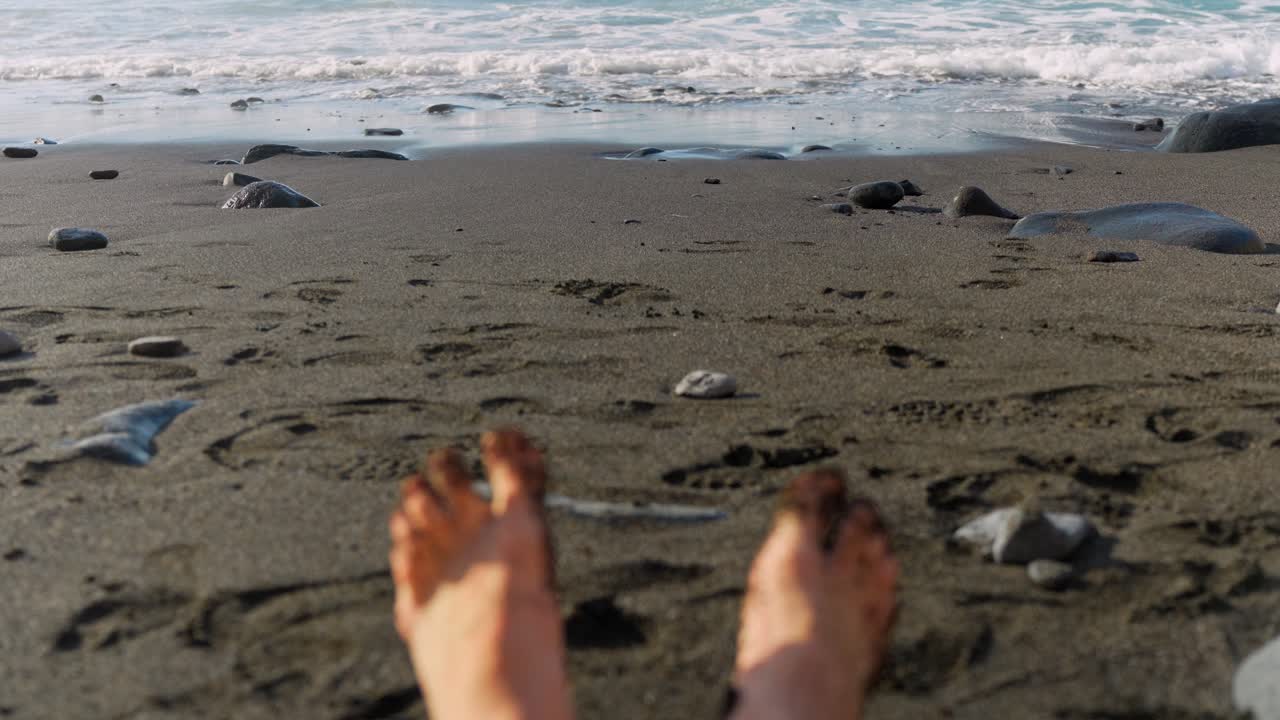 los dedos de los pies de arena negra se mueven en cámara lenta mientras las olas del océano chocan contra la playa en cámara lenta