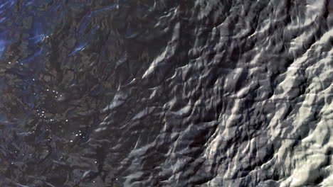 overhead drone frames dark water sculpted by wind into ripples and pale foam, an abstract surface of shifting light and texture that suggests calm motion and meditative focus