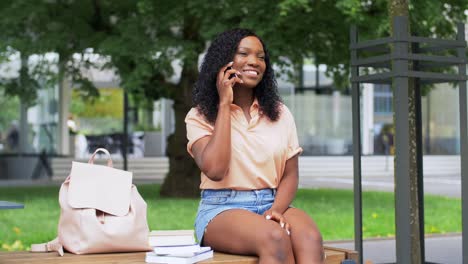 Concepto-De-Tecnología,-Educación-Y-Personas-Feliz-Y-Sonriente-Estudiante-Afroamericana-Con-Libros-De-Texto-Llamando-A-Un-Teléfono-Inteligente-En-La-Ciudad