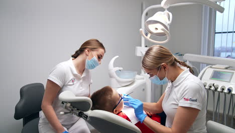 lady dentist in white uniform applying metal instruments in her work. professional is examining the little boy sitting in dental chair.