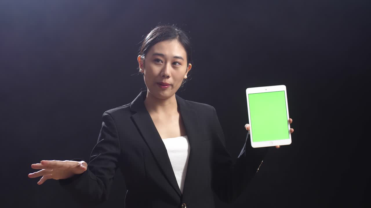 블랙 스크린 스튜디오 에서 연설 하는 동안 녹색 스크린 태블릿 을 들고 가리키는 비즈니스 슈트 를 입은 아시아 여성