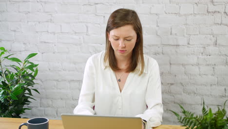 A-young-woman-working-on-a-laptop-comuter-in-a-white-walled-office-with-plants-and-a-coffee-cup