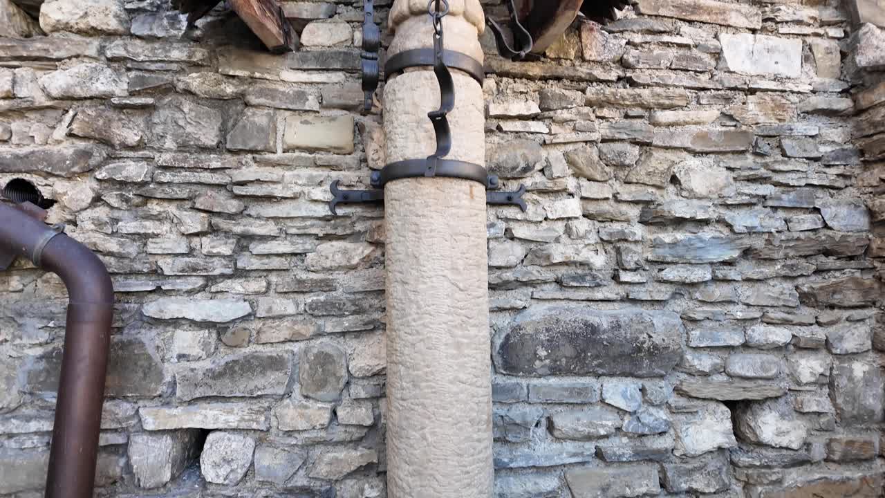 White stone pillory with black iron shackles mounted to castle walls. A heavy chained sphere lies below. Historic Slovak setting evoking medieval justice, punishment, and architectural heritage