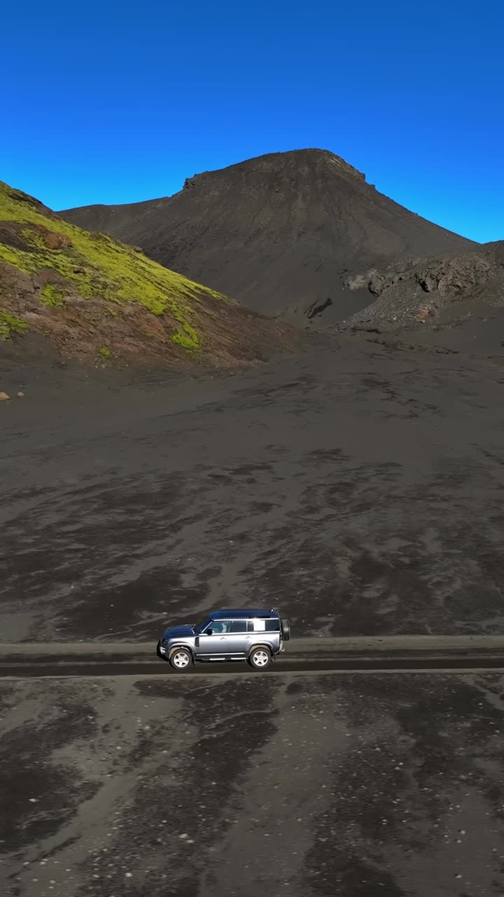 Vista aerea verticale da drone di un SUV 4x4 argento che attraversa una pianura islandese di sabbia nera sotto un cielo azzurro, con una robusta collina vulcanica sullo sfondo