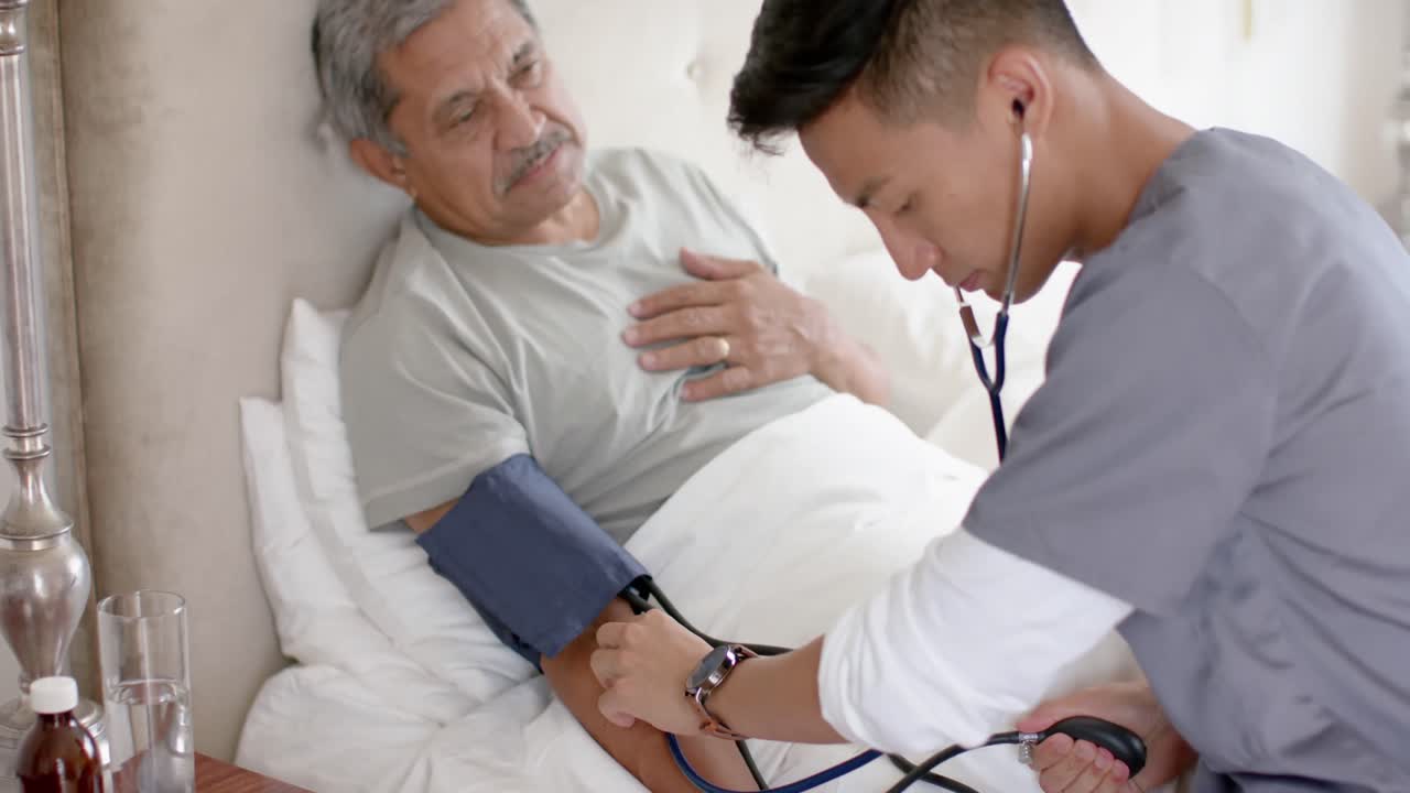 Medico uomo multietnico che misura la pressione sanguigna a paziente anziano uomo a letto a casa, al rallentatore