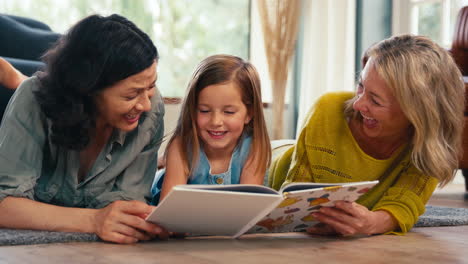 familia del mismo sexo con dos madres e hija acostadas en el suelo leyendo un libro juntos en casa