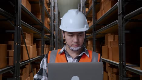 close up of asian male engineer with safety helmet working on a laptop while standing in the warehouse with shelves full of delivery goods