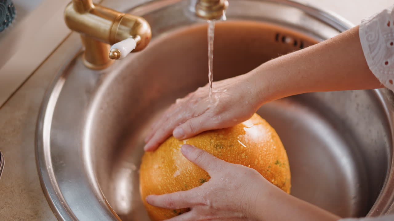 Manos blancas enjuagan una calabaza bajo el grifo, en un fregadero de acero inoxidable con un chorro de agua constante. Un primer plano muestra la cáscara naranja texturizada y las gotas brillantes, reflejando una preparación metódica y limpia por parte de un cocinero casero para un pastel.