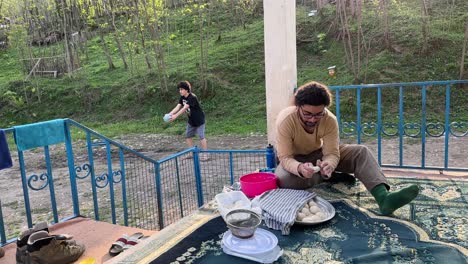 a man is kneading bread dough to bake flat bread in clay oven while a young boy is playing basketball with a plastic ball in the yard in rural village area in middle east asia