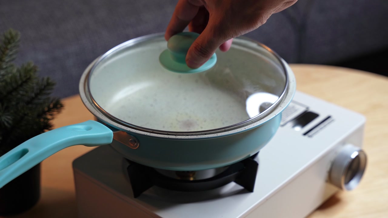shot of a person's hand placing a glass lid on a light blue speckled frying pan. The empty cookware sits on a white portable gas stove on a wooden table in a modern indoor home setting.