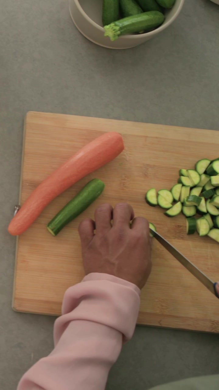 Video verticale: Una mano con manica rosa entra in scena, affettando zucchine a rondelle su un tagliere a casa, con un coltello per la preparazione.