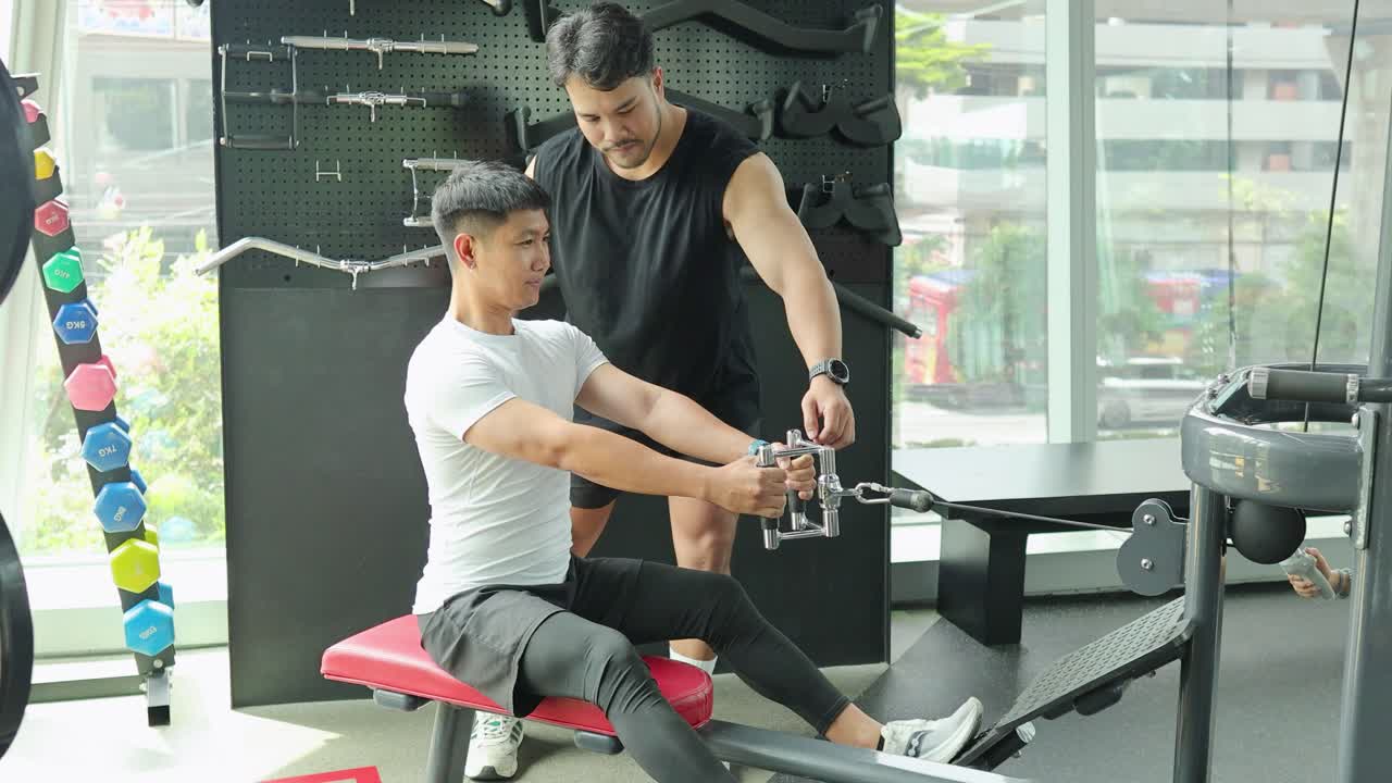 A man exercises on a cable machine under guidance in a bright gym setting