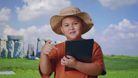 asian boy with a hat and binoculars smiling and pointing to a tablet while traveling in stonehenge. boy researcher examines something, travel tourism adventure concept, close up