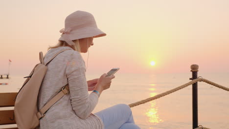 una chica elegante con ropa rosa escuchando música en un teléfono inteligente y sentada en un banco en un mar pi