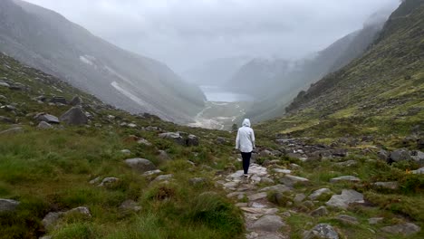 Vista-Trasera-De-Una-Persona-Femenina-Con-Impermeable-Caminando-Por-Un-Camino-Rocoso-Entre-Montañas-Gigantes-De-Irlanda-Durante-Un-Día-Nublado-En-La-Naturaleza---Toma-Amplia-En-Cámara-Lenta