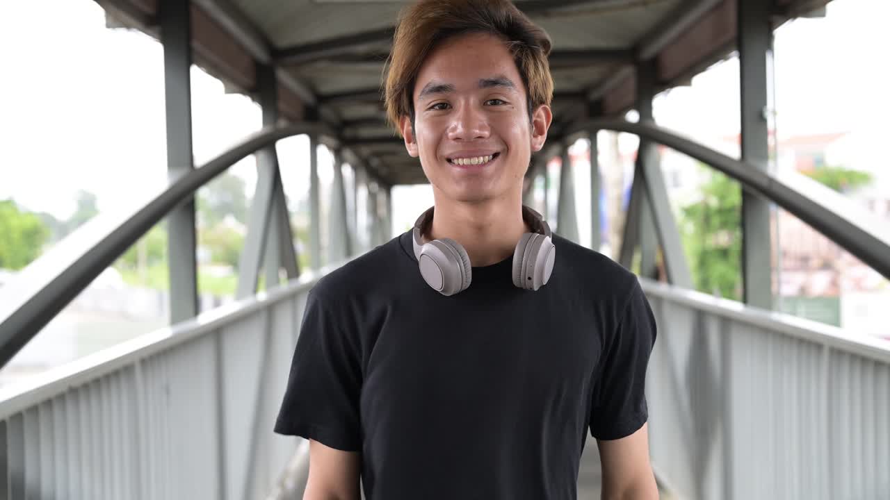 Portrait of a young Thai Asian man in casual style wearing black t-shirt and headphones looking at camera and smiling in walking bridge during windy summer day