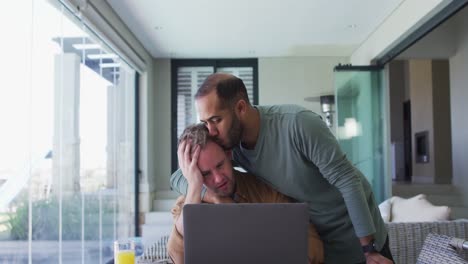 pareja multiétnica masculina del mismo sexo sentada en la mesa en casa usando una computadora portátil y abrazándose