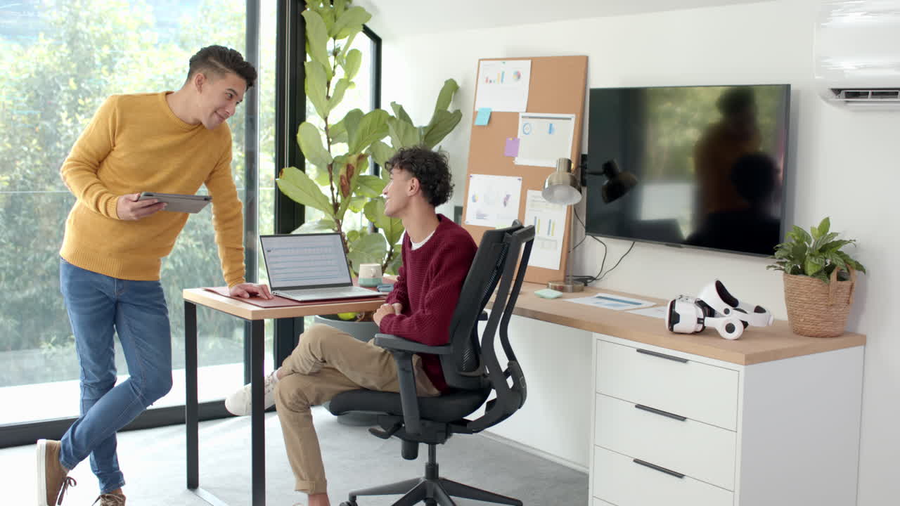 Discussing work, multiracial gay couple using laptop and tablet in modern home office