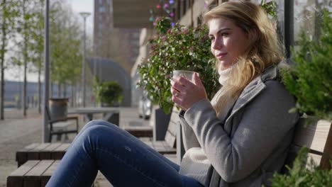 Attractive-woman-sitting-on-a-terrace-in-the-sun,-blowing-a-hot-beverage-and-taking-a-sip,-panning-slow-motion