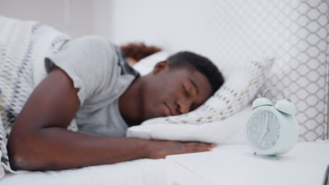 man asleep in bed reaches out to turn off alarm clock on bedside table