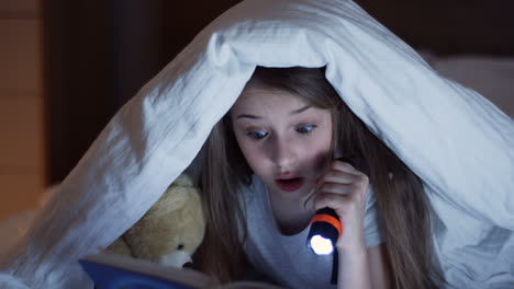 close-up view of cute little girl using a flashlight and reading a book with surprised expression under the blanket at night