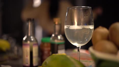 single young woman dancing in her modern kitchen while cooking dinner for one - focus on single wine glass with her reflection in foreground and out of focus background