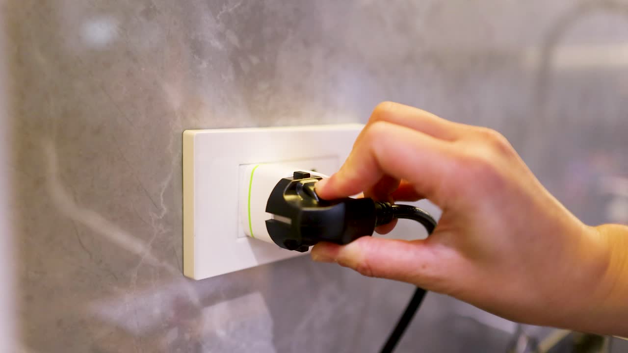 A person's hand connects a black electrical plug into a white European wall outlet