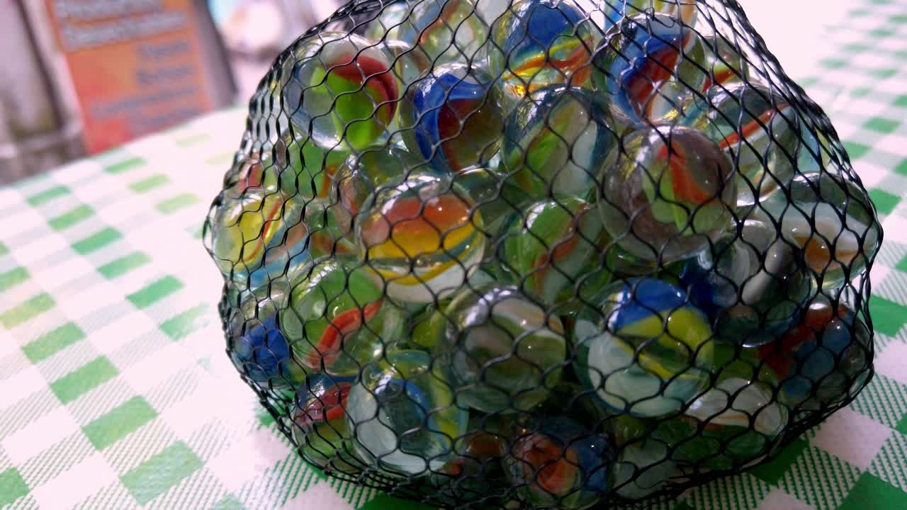 Rotating close-up shot of colorful glass marbles inside a mesh bag placed on a green checkered tablecloth