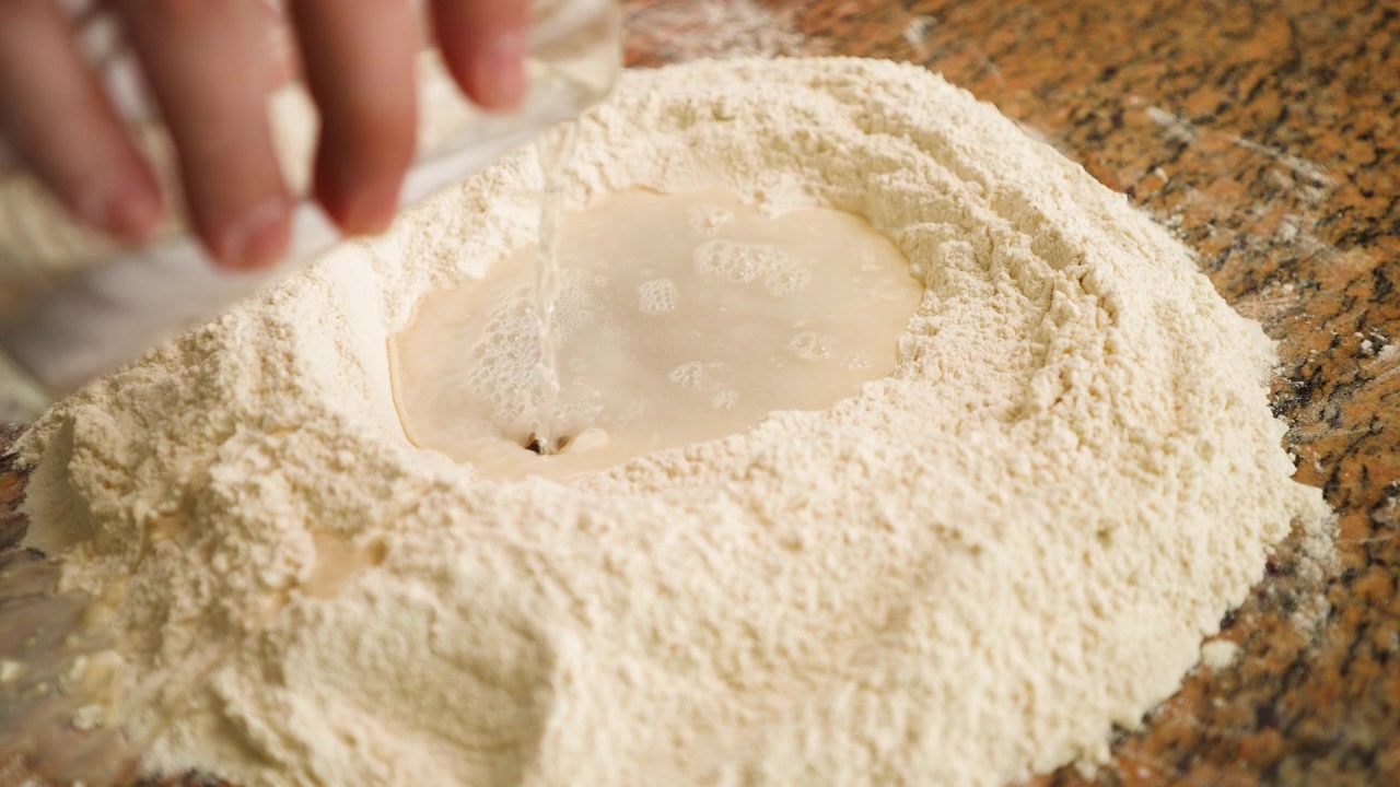 Captivating footage of water streaming into flour, signaling the beginning of a traditional pizza dough recipe