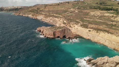 Aerial-POI-drone-shot-captures-the-rugged-rock-formations-along-Gozo's-coast,-moving-from-left-to-right,-showcasing-the-stunning-turquoise-sea,-with-Mgarr-city-and-port-in-the-background