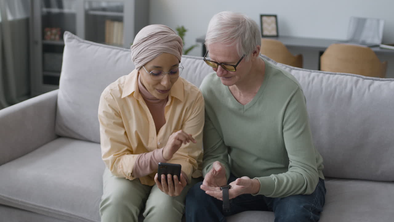 Middle Aged Arabic Woman Helping A Senior Man To Sync A Smartwatch To Phone While Sitting On Sofa At Home
