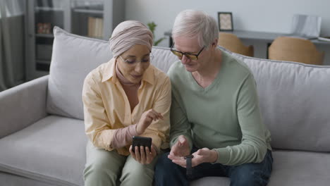 Mujer-árabe-De-Mediana-Edad-Ayudando-A-Un-Anciano-A-Sincronizar-Un-Reloj-Inteligente-Con-El-Teléfono-Mientras-Se-Sienta-En-Un-Sofá-En-Casa