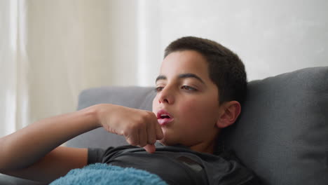 little kid with brown eyeball looks visibly sick and exhausted as he lies on couch near window coughing and adjusting his position, expressing flu symptoms and discomfort in calm home environment
