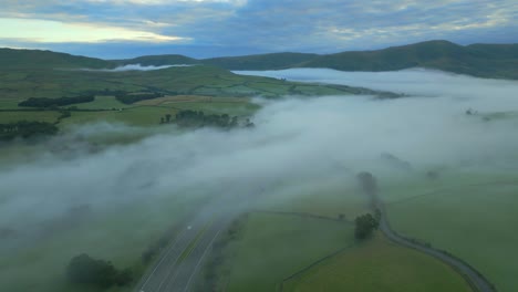 Flying-high-over-M6-motorway-with-slow-pan-revealing-misty-countryside,-fog-in-valley,-green-fields-and-hills,-mountains-at-sunrise