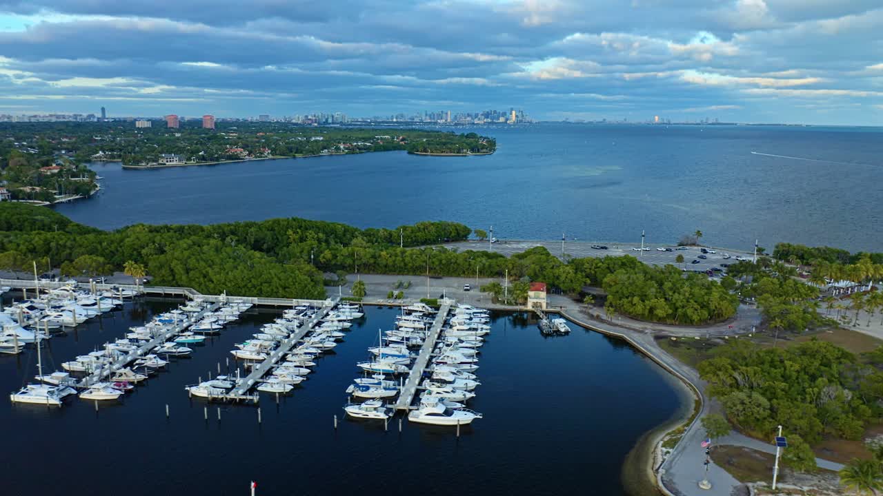 Rows of sailboats fill the marina beside dense mangroves as Biscayne Bay stretches toward the distant Miami skyline, forming a wide coastal scene at Matheson Hammock Park