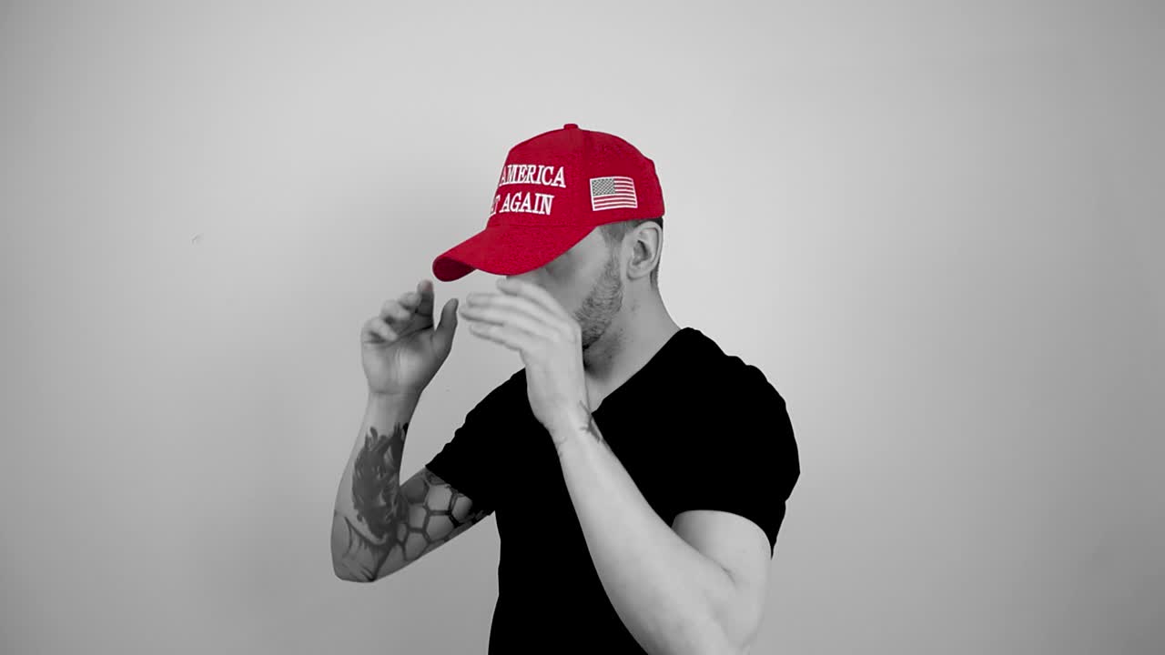 Grayscale footage of a tattooed man with short hair and bear putting on a red vibrant colored MAGA Make America Great Again Hat, then looking forward into camera and doing thumbs up with lifting arms.