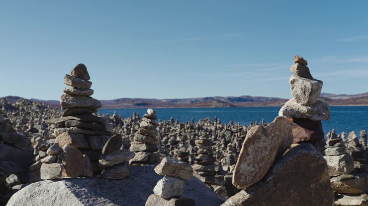 Stakke af tørre sten danner tætte vardeklynger ved et bredt reservoir på Hardangervidda i skarpt middagslys, med fjerne afrundede bakker bag vandet