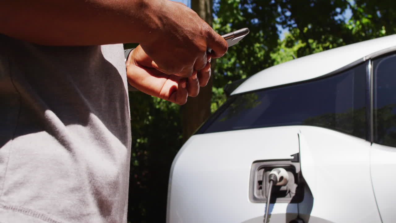 sección media de un hombre mayor afroamericano cargando un coche eléctrico y usando un teléfono inteligente en un día soleado