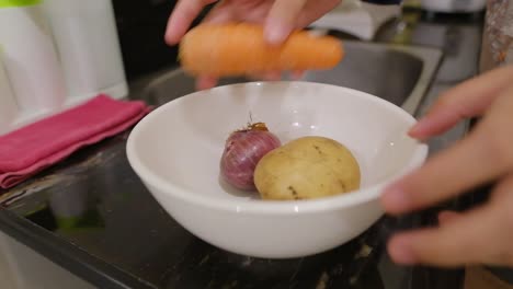 person places fresh raw potato, red onion, and carrot in white ceramic bowl for washing in the kitchen sink