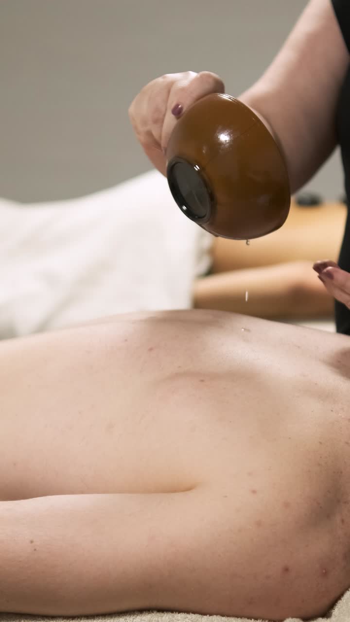 Masseuse skillfully pouring aromatic oil onto a client's back for a soothing massage. Vertical