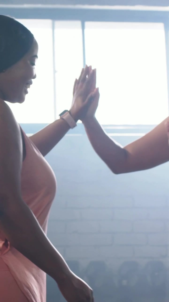 Vertical video: Pal entering urging her in pink tank rising high-fiving at gym weights, copy space