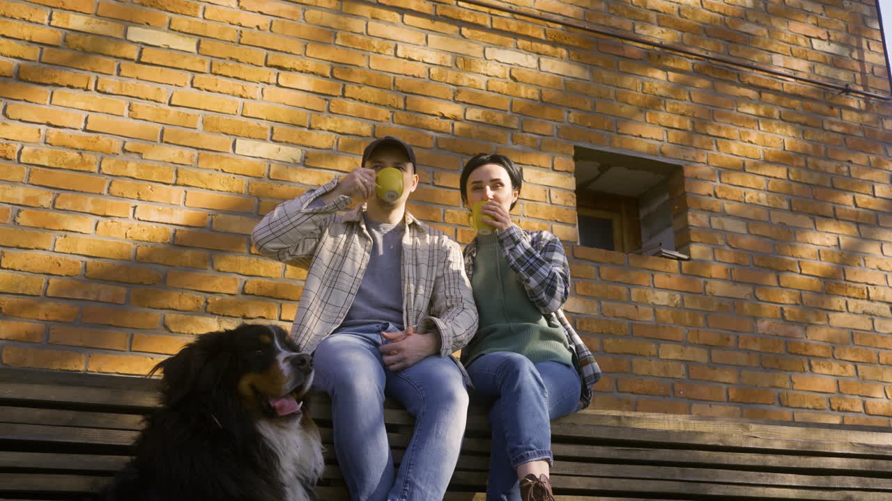 백인 부부가 시골 집 밖에서 차를 마시면서 대화를 나누는 바닥 시야. 그들의 개는 그들 근처에 땅에 누워 있습니다.