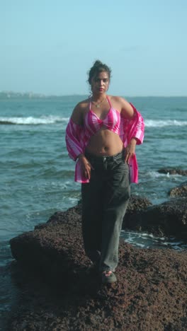 young woman standing on rocks by the ocean, wearing a pink top and loose jeans