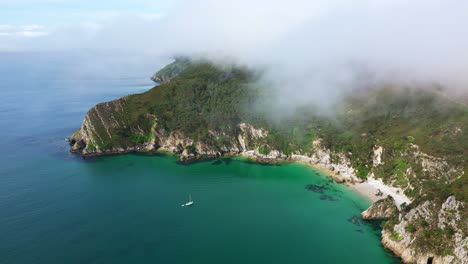 drone panning above île vierge beach in crozon peninsula, brittany, showing cliffs, turquoise sea, sailboat, coastal vegetation, and light mist - france